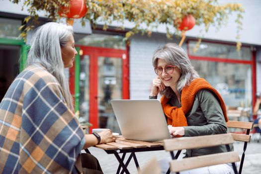 Hoary haired Asian woman and mature friend with laptop sit together at small table on outdoors cafe terrace on nice autumn day