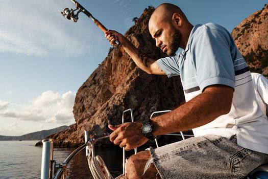 Young african american man standing with fishing rod on a sailboat fishing in open sea on sunset, close up