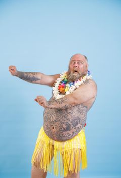 Emotional bearded man with overweight in decorative yellow grass skirt and flowers garland dances on light blue background in studio