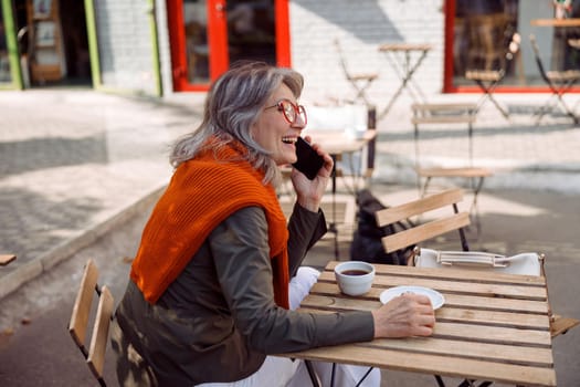 Positive grey haired senior lady with glasses and sweater talks on mobile phone at table on outdoors cafe terrace on autumn day