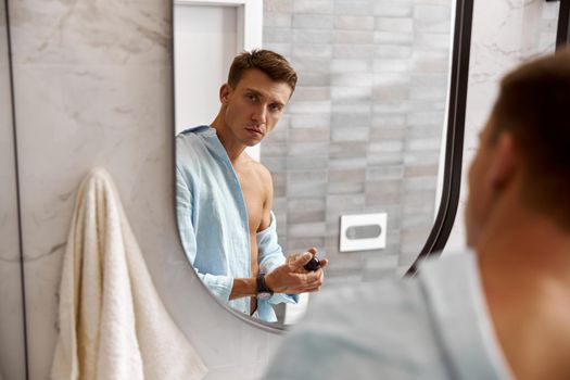 Photo of adult caucasian beautiful man in front of mirror in light modern bathroom