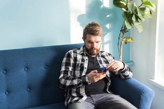 Technologies and leisure concept - Cheerful man sitting on the couch using his smartphone at home in living room.