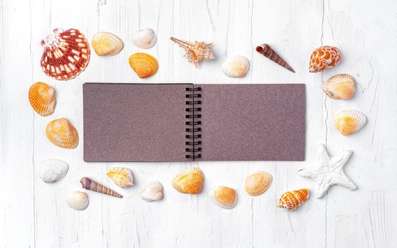 Open notepad with sea shells on white wooden background, top view