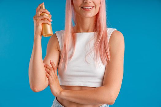 Cropped shot of young woman with pink hair smiling and holding bottle of moisturizing spray while standing isolated over blue studio background. Beauty products, hair care concept