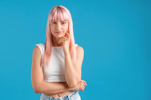 Beautiful young woman with pink hair looking at camera, standing isolated over blue studio background. Emotions, lifestyle concept