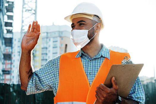 Portrait of mixed race man builder in workwear and hardhat wearing medical mask, close up photo