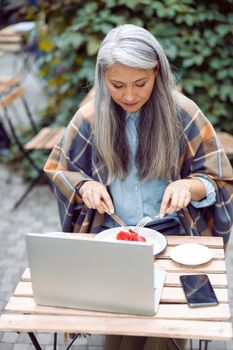 Pretty long haired senior Asian woman eats toast with fresh strawberries in front of open laptop at table on outdoors cafe terrace on autumn day