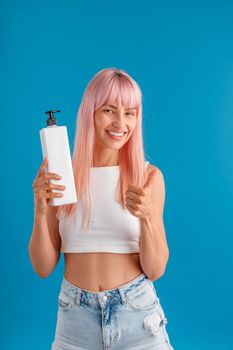 Smiling woman with smooth pink hair showing thumbs up and holding pump bottle with hair mask or shampoo, standing isolated over blue studio background. Beauty and hair care concept