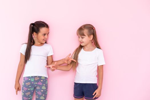 Amazed little kids girls showing white t-shirt, isolated on pink background. Childhood lifestyle concept. Mock up copy space. Pointing index finger aside