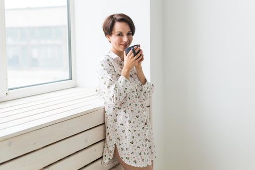 Beautiful young woman with cup of tea standing near the window at home