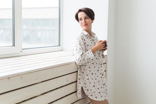 Beautiful young woman with cup of tea standing near the window at home