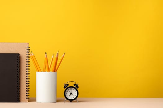 Cup of pencils and notepads on desk against yellow background, front view