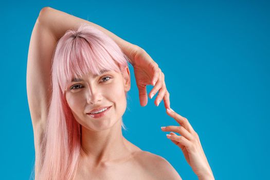 Portrait of naked tattooed woman with pink hair and perfect skin smiling at camera, posing with raised hands isolated over blue studio background. Beauty, skin care concept