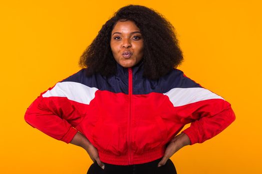 Funny curly african american girl dressed in the red sports jacket in the studio on yellow background. Fashion and people.