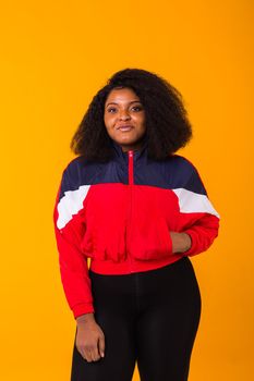 Funny curly african american girl dressed in the red sports jacket in the studio on yellow background. Fashion and people.