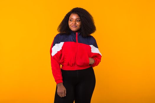 Funny curly african american girl dressed in the red sports jacket in the studio on yellow background. Fashion and people.