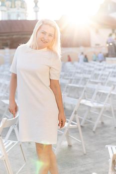 attractive woman dressed in white dress in summer open air theater on chair alone, spring street style fashion trend, social distancing