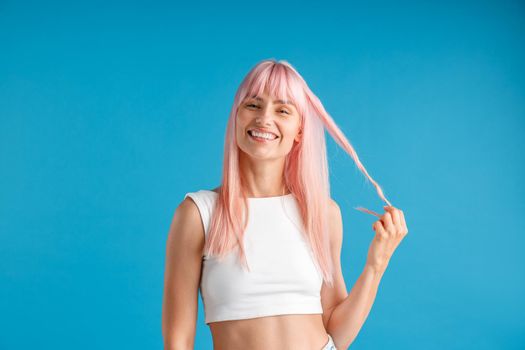 Pleased young woman with natural long pink dyed hair holding a strand of it and smiling at camera, posing isolated over blue studio background. Beauty, hair care concept
