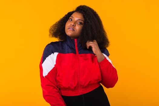 Funny curly african american girl dressed in the red sports jacket in the studio on yellow background. Fashion and people.