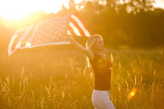 Beautiful young girl holding an American flag in the wind in a field of rye. Summer landscape against the blue sky. Horizontal orientation.