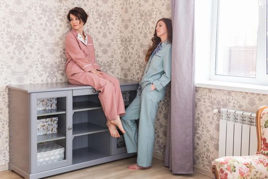Two female sisters or friends relaxing at the living room. Girls in stylish pajamas. Quarantine and stay at home.