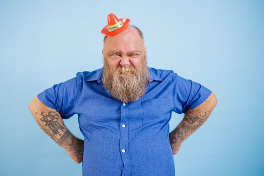 Funny bearded plus size man wearing small sombrero and tight blue shirt holds hands on waist grimacing on light blue background in studio