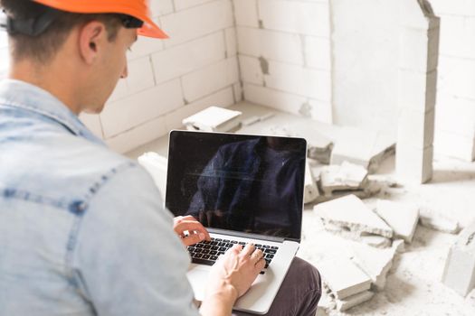 Handsome architect or foreman in helmet working with laptop on the apartment drawings at the structure white interior