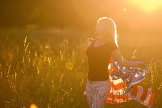 Beautiful young girl holding an American flag in the wind in a field of rye. Summer landscape against the blue sky. Horizontal orientation.