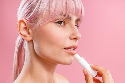 Close up portrait of beautiful young woman with pink hair looking aside, applying lip balm on her lips isolated over pink background. Beauty, lip care concept