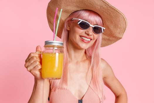 Portrait of happy woman wearing straw hat, swimsuit and sunglasses smiling at camera, drinking tropical juice cocktail, posing isolated over pink studio background. Summer vacation concept