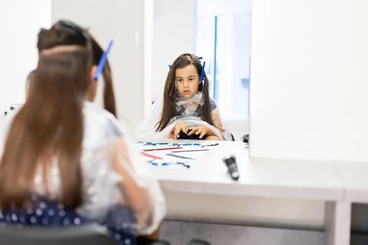 Portrait of pretty little girl seating at table with accessories in a hairdressing salon