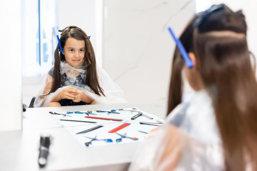 Portrait of pretty little girl seating at table with accessories in a hairdressing salon