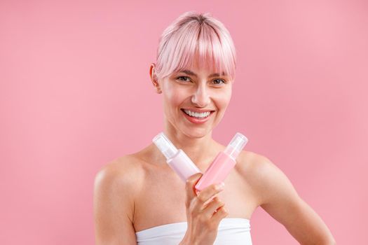 Excited young woman with pink hair smiling and showing two plastic spay bottles with beauty products, posing isolated over pink background. Beauty, spa, body care concept