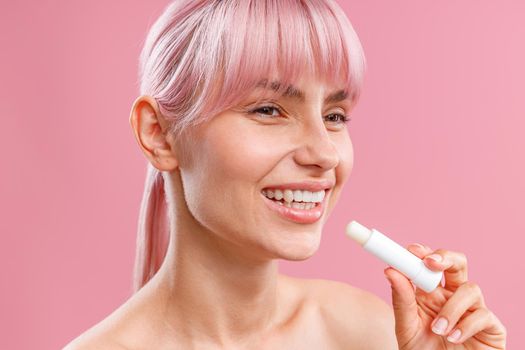 Close up portrait of cute young woman with pink hair smiling aside, holding lip balm near her lips isolated over pink background. Beauty, lip care concept