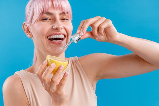 Portrait of joyful woman with perfect skin and pink hair laughing, holding half of fresh lemon and a dropper isolated over blue background. Vitamin C serum. Beauty, health concept