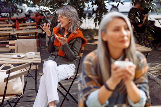 Senior cafe guests, focus on happy silver haired woman with glasses recording audio message on mobile phone on outdoors cafe terrace