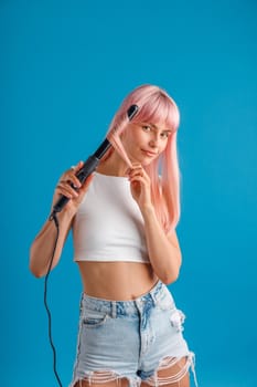 Attractive young woman looking at camera, curling pink long straight hair using curling iron, standing isolated over blue studio background. Beauty and hair care concept