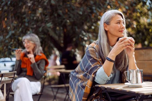 Senior cafe guests, focus on thoughtful hoary haired mature woman holding cup at table on outdoors cafe terrace on autumn day