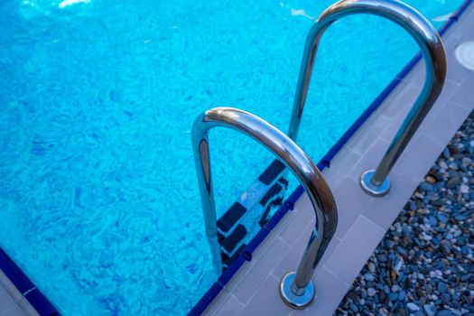 Background of blue transparent water in the swimming pool on the territory of the hotel. Descent to the pool with handrails. Rest and relaxation concept. Active rest by the sea.