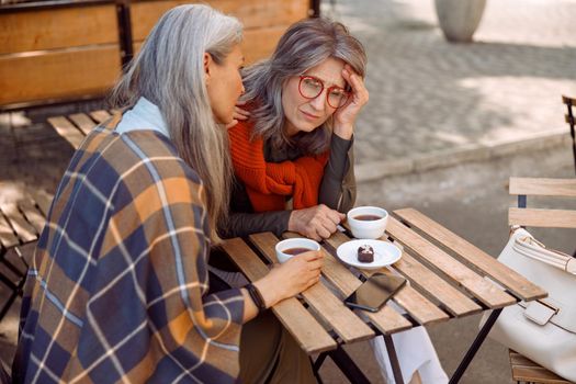 Depressed mature lady with glasses and careful friend sit together at small table in street cafe on nice autumn day