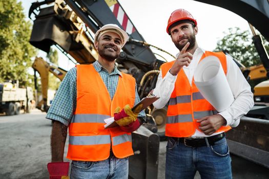 Two men engineers in workwear discussing their work standing against construction machines