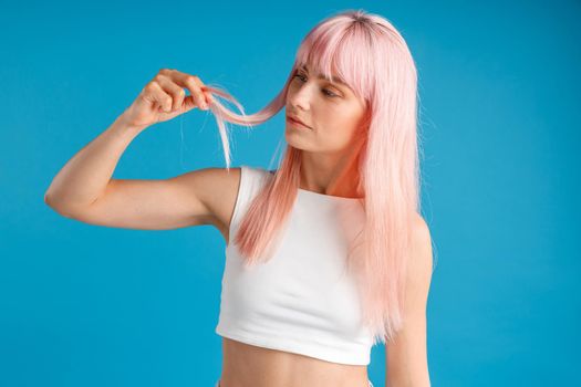 Curious woman touching and looking at her smooth natural long pink dyed hair while posing isolated over blue studio background. Beauty, hair care concept