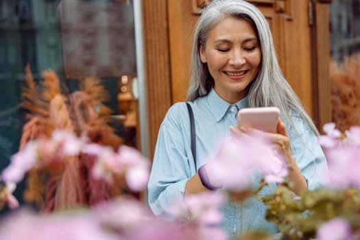 Beautiful silver haired mature Asian woman in denim shirt uses mobile phone on outdoors terrace decorated with flowers