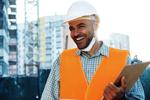 Portrait of mixed race man builder in workwear and hardhat wearing medical mask, close up photo