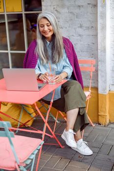 Smiling senior Asian woman with silver hair and glass of water sits at coral table with open laptop on outdoors cafe terrace