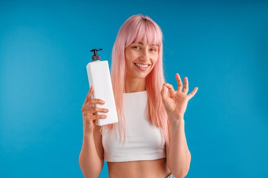 Smiling woman with smooth pink hair showing ok sign and holding pump bottle with hair mask or shampoo, standing isolated over blue studio background. Beauty and hair care concept