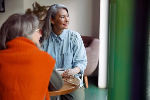 Cheerful senior woman with friend look out of window sitting at small table in cozy cafe. Long-time friendship