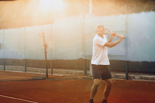 Young man plays tennis outdoors on clay tennis court in the morning