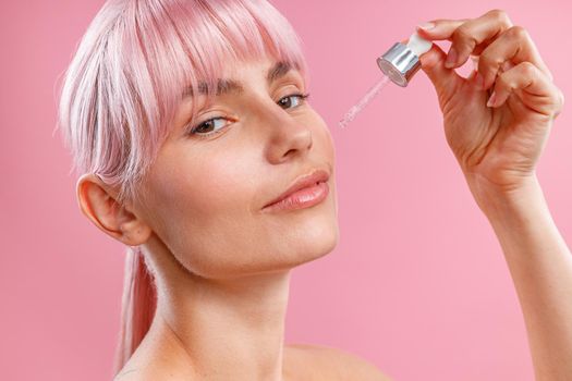 Close up portrait of young woman with pink hair applying serum or hyaluronic acid on her facial skin using dropper isolated over pink background. Beauty, cosmetics and skincare concept