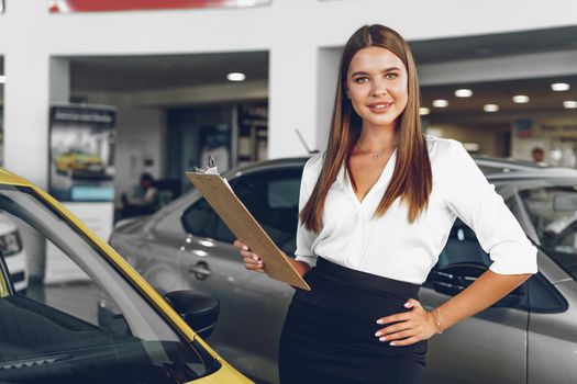 Attractive young female car dealer standing in showroom near a new car